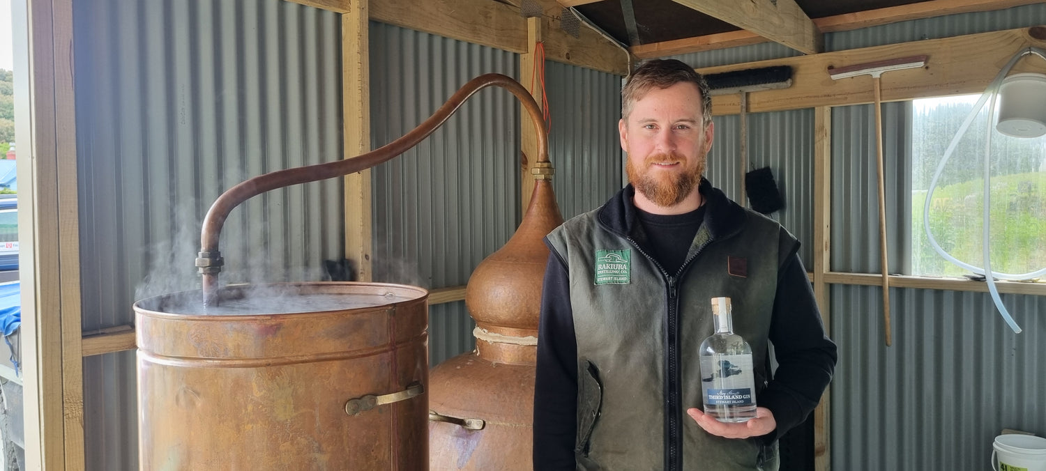Man holding a bottle of Third Island Navy Strength Gin next to a large copper still in a rustic setting.