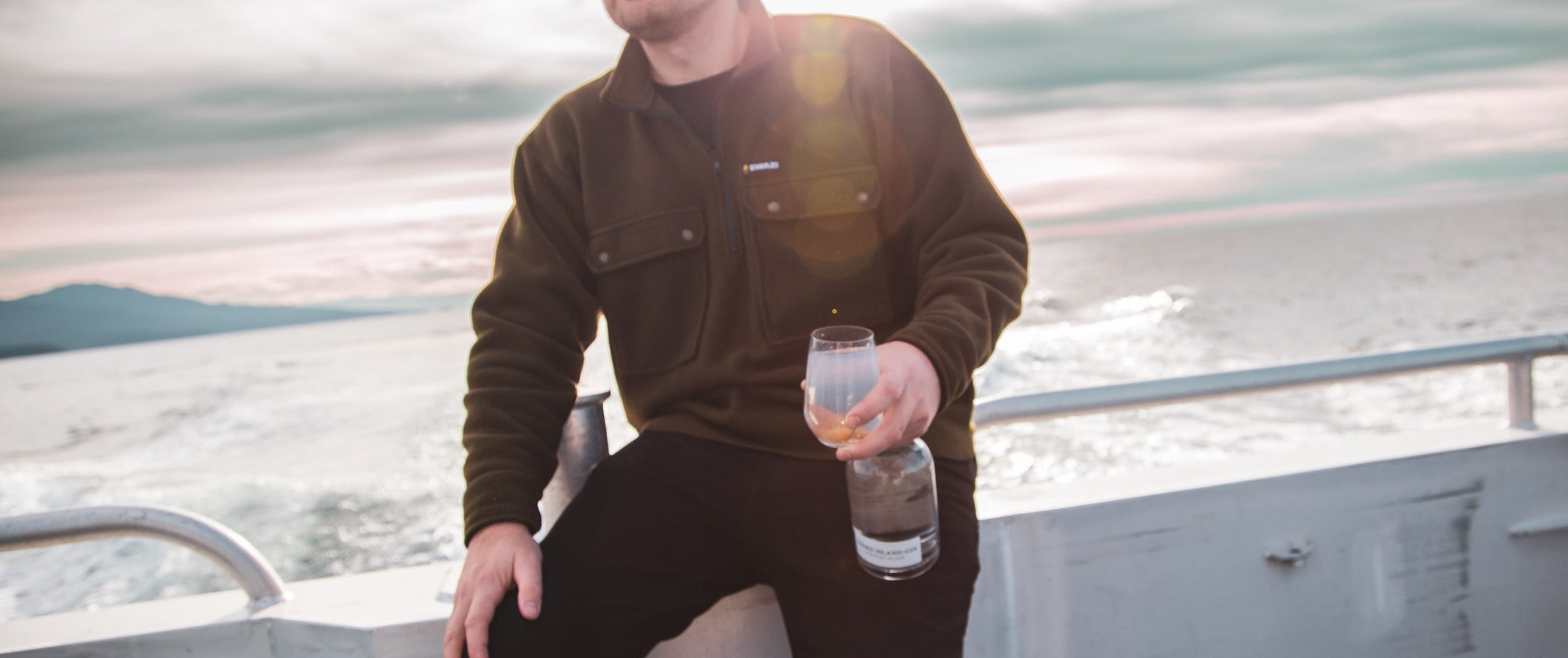 Person sitting on a boat holding a bottle and glass of Third Island Gin with a scenic background