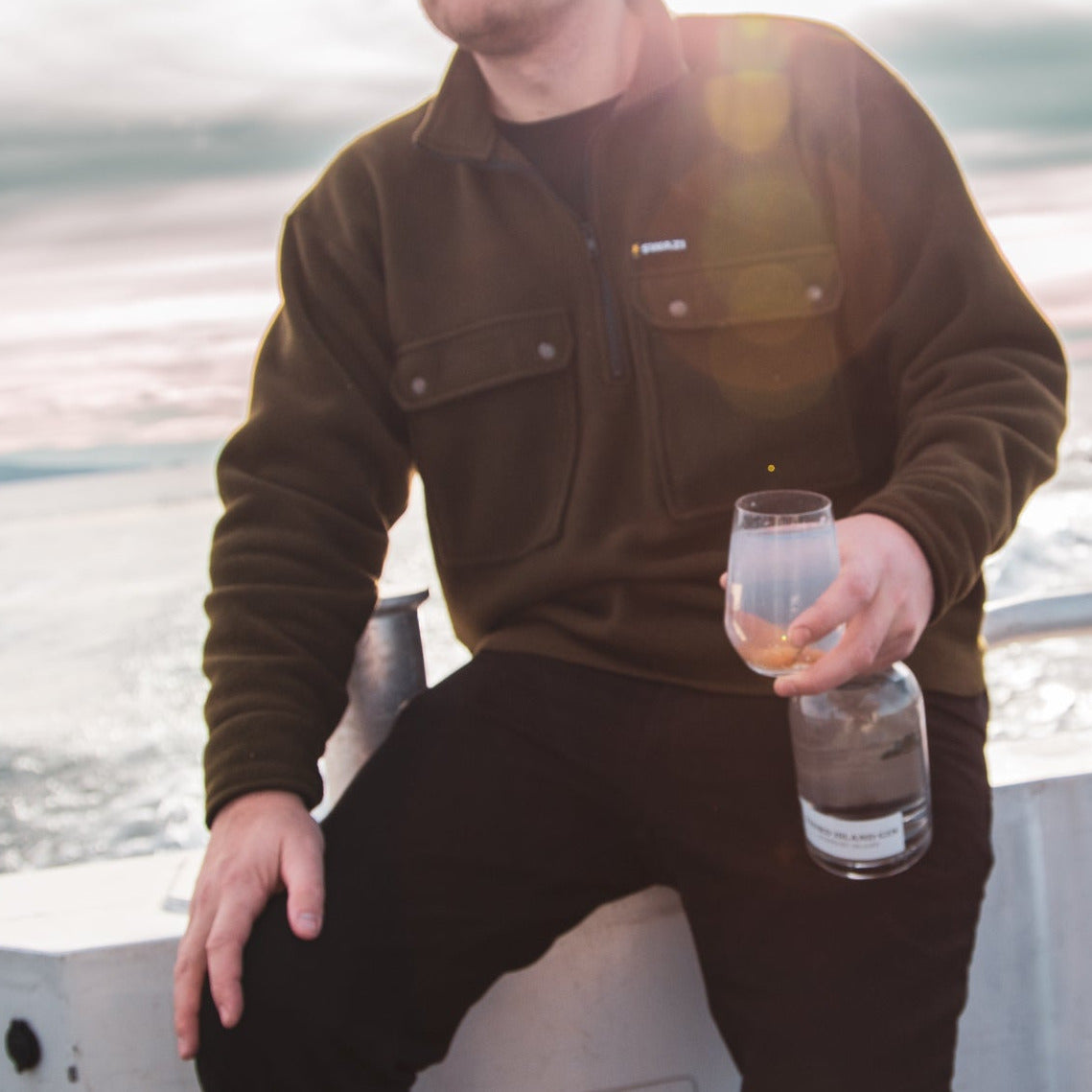 Person sitting by a body of water holding a glass and bottle of Third Island Gin
